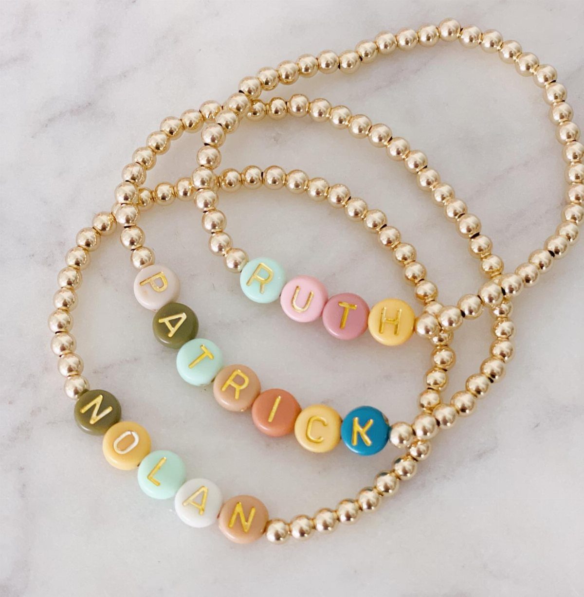 11 Beaded Jewelry DIY Bracelets Ideas for Every Style 5 Three gold beaded bracelets with colorful alphabet beads spelling Ruth, Patrick, and Nolan.
