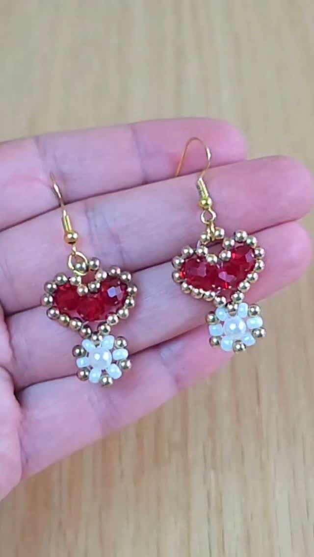 Beaded heart earrings with red centers and white flower accents held in a hand.
