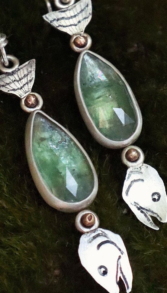 Silver earrings with green teardrop gemstones and fish charms on green moss.