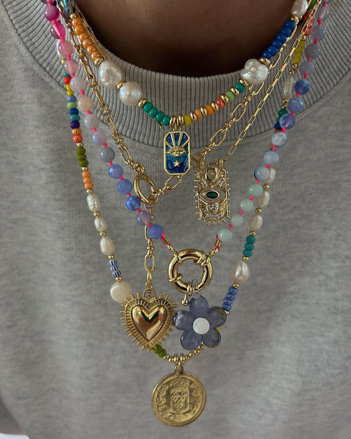 Layered colorful beaded necklaces with gold charms worn over a grey ribbed shirt.