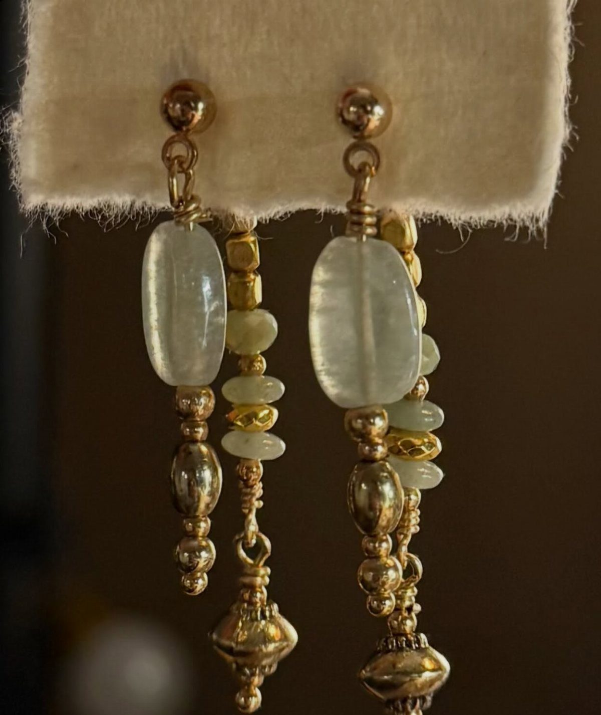 Gold-toned earrings with pale green beads displayed on a beige fabric stand.