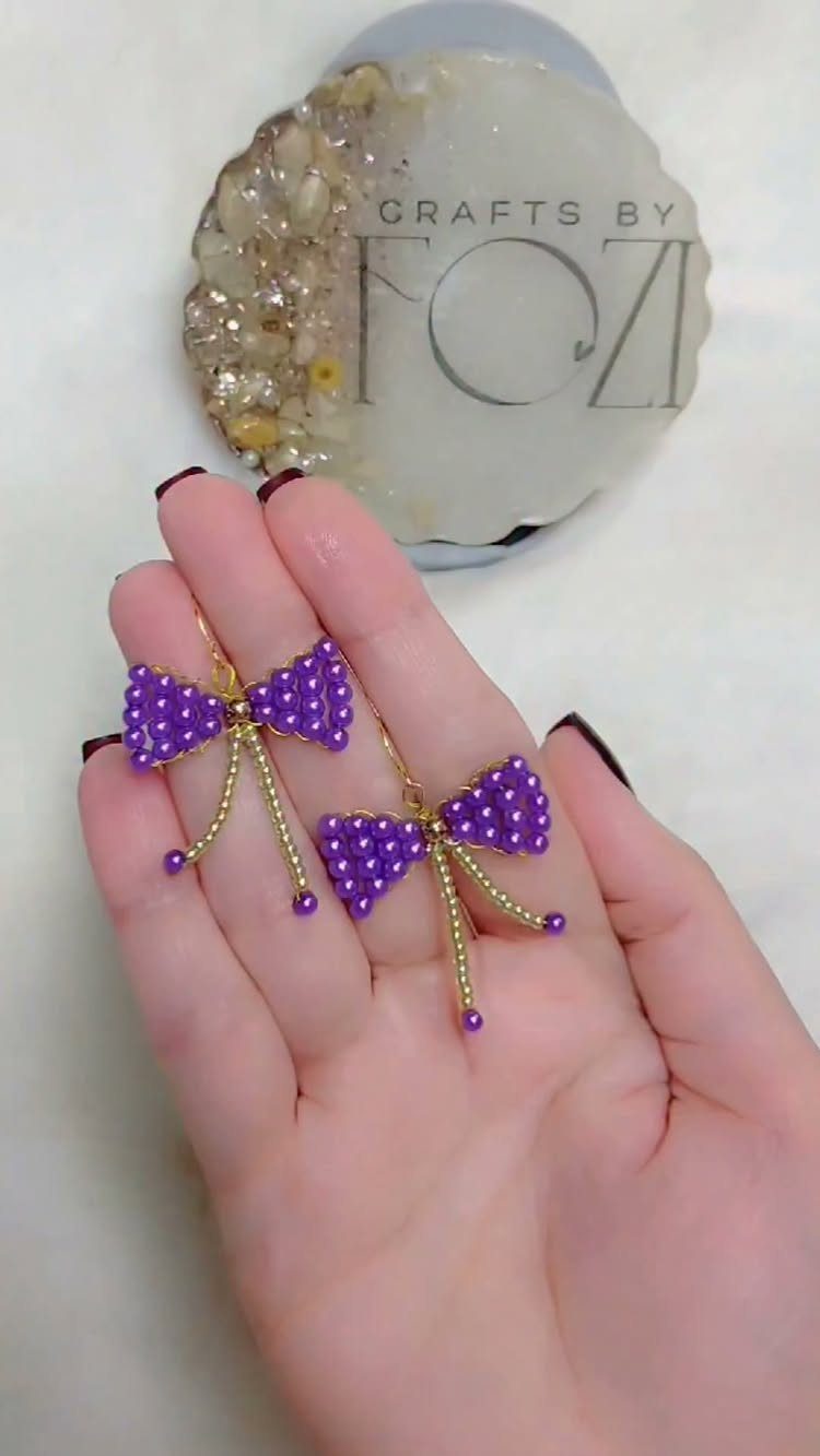 Purple beaded bow earrings held in a hand with a craft coaster background