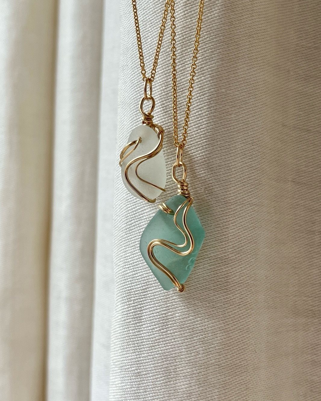 Two sea glass pendants wrapped in gold wire hanging against white fabric