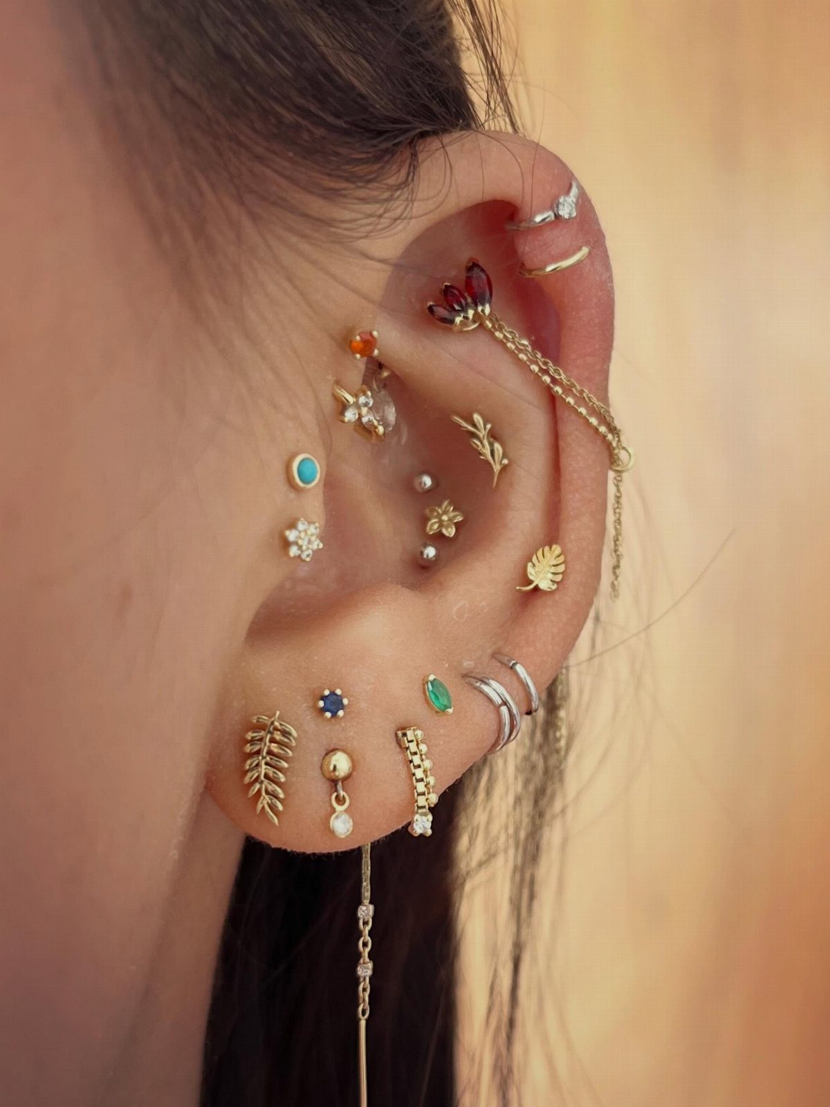 Ear with multiple gold piercings featuring floral and leaf-shaped studs and colorful gemstones.