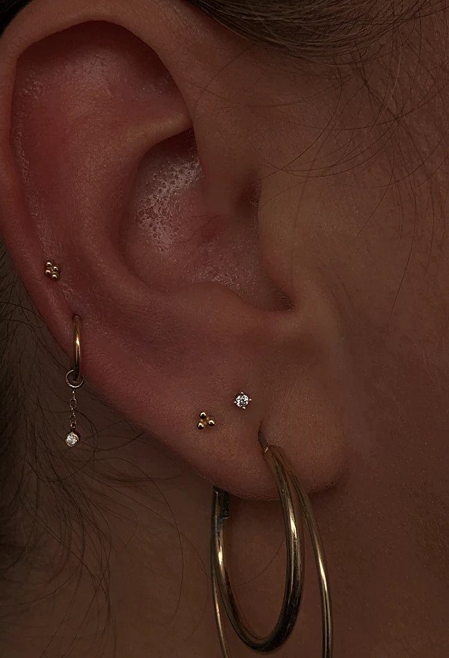 Ear with multiple gold piercings including tiny studs and gold hoop earrings.