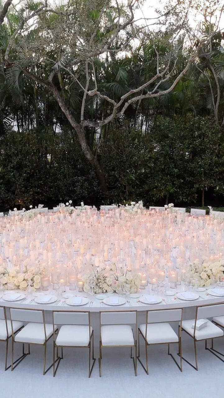 White curved dining table surrounded by hundreds of glowing candles and white flowers outdoors.