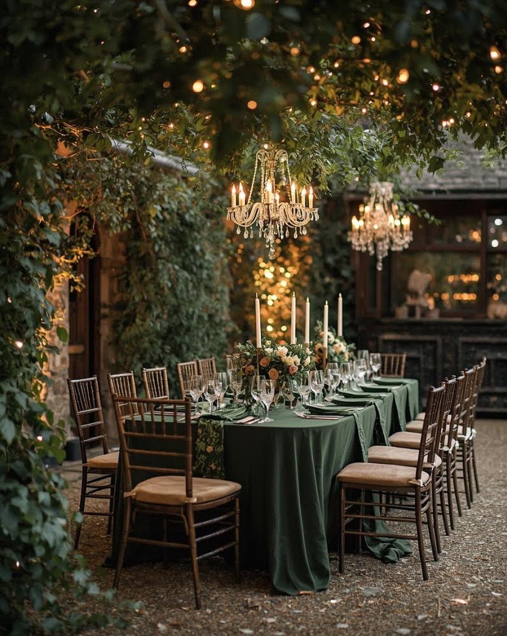 Outdoor dining table with green linens and crystal chandeliers under a leafy canopy.