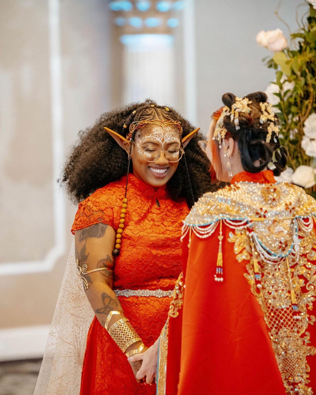 Two people in red fantasy costumes with gold details and elf ears facing each other.