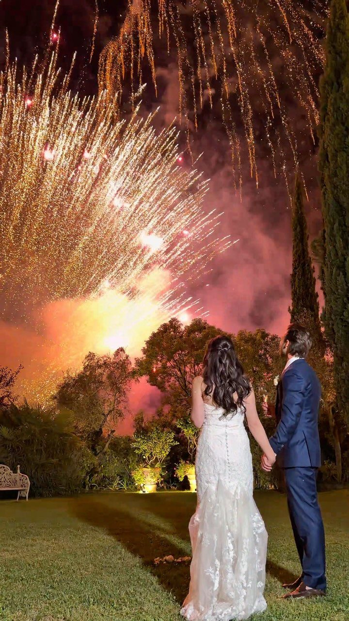 Newlyweds holding hands while watching a golden fireworks display in the night sky.