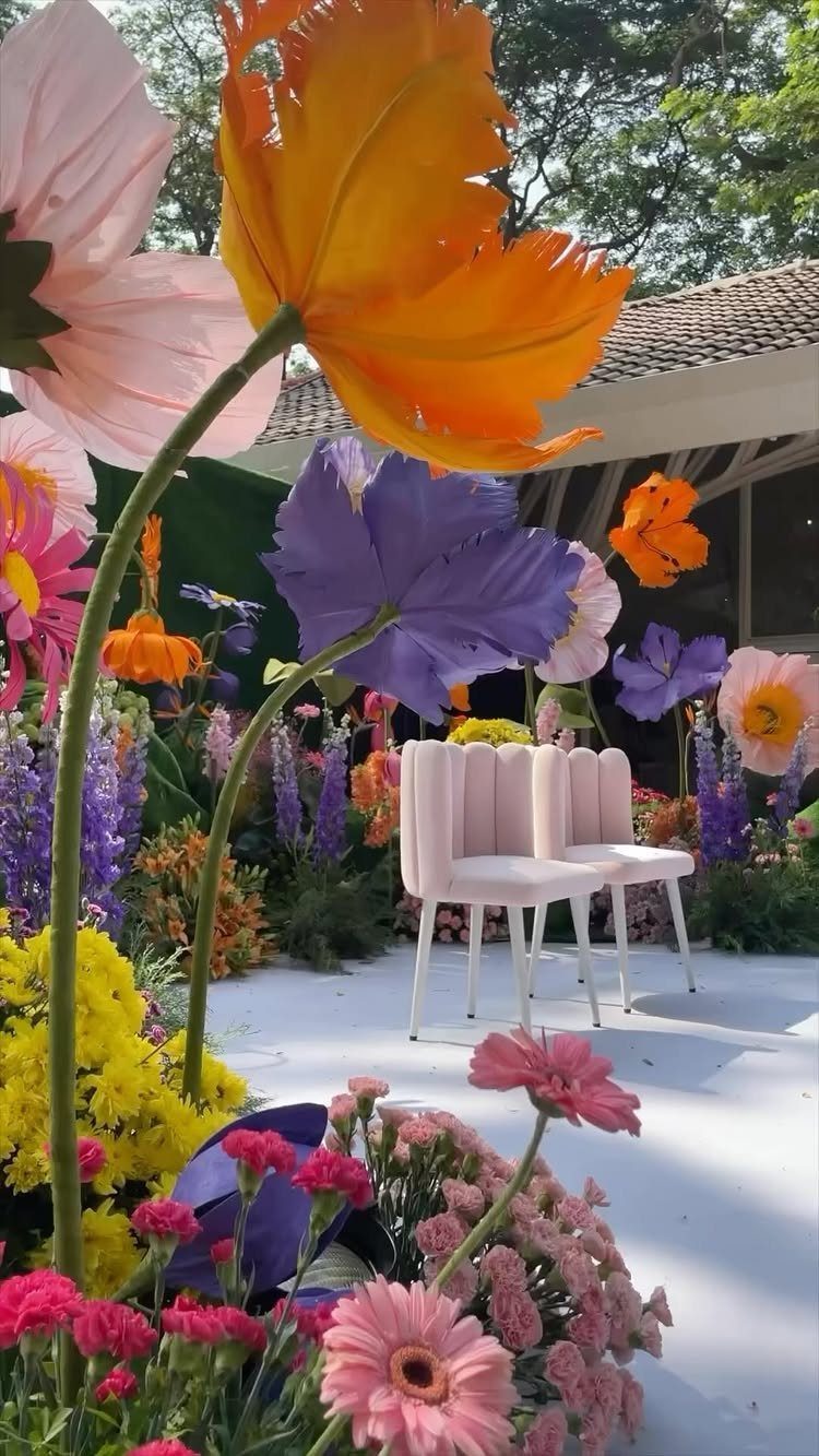 Giant colorful artificial flowers surrounding pale pink chairs in a whimsical outdoor setting