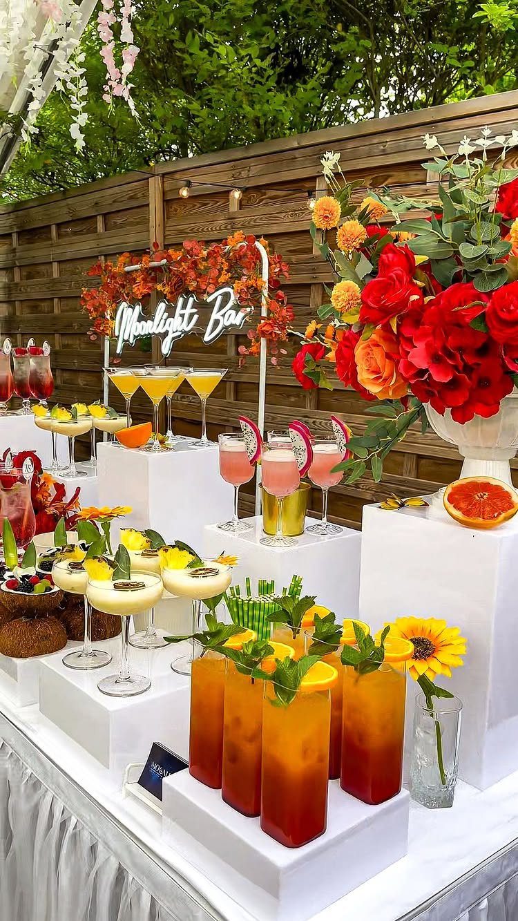 Colorful cocktails on white tiered pedestals at an outdoor wedding bar with flowers.