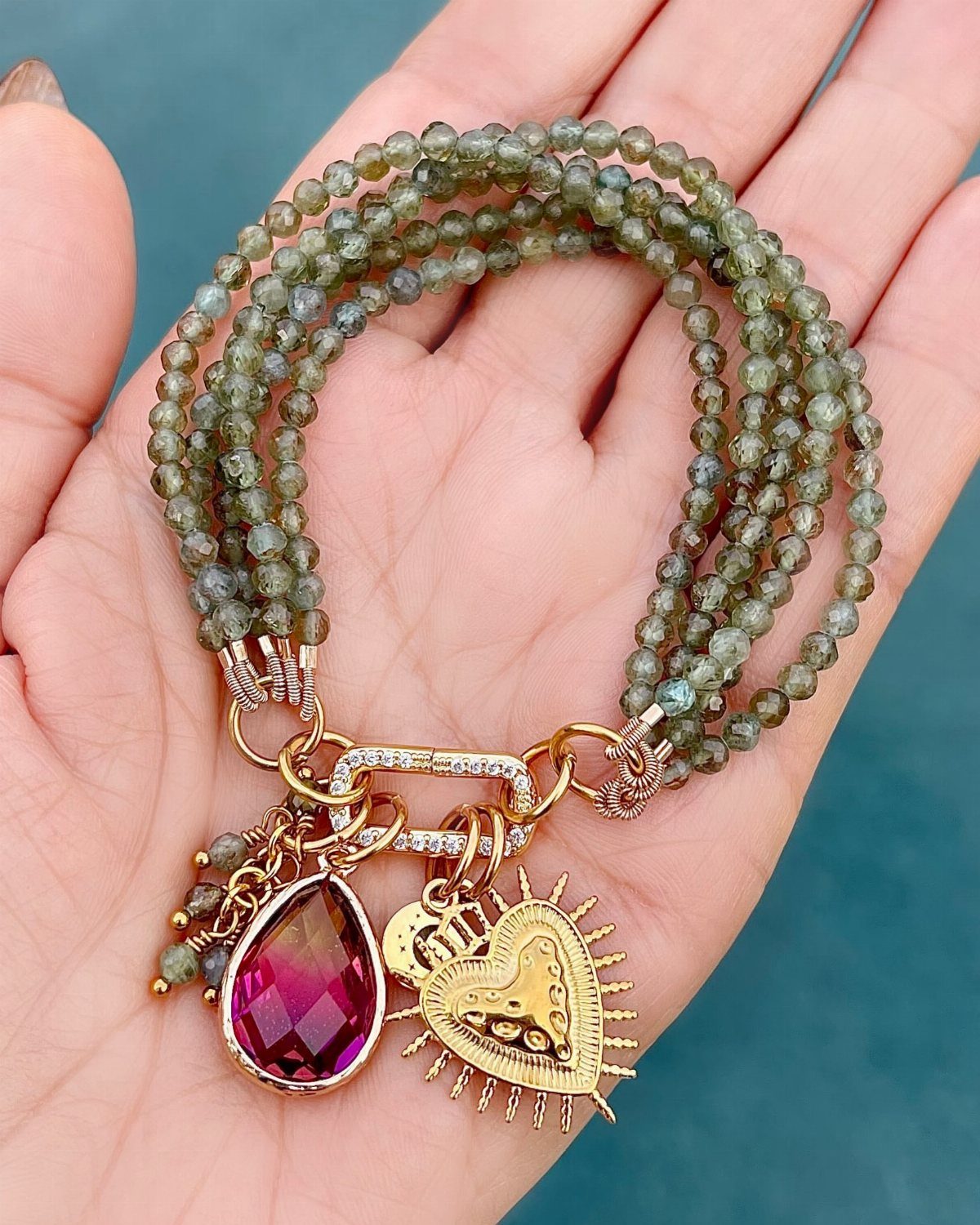 Multi-strand green beaded bracelet with gold heart and purple gem charms in a palm