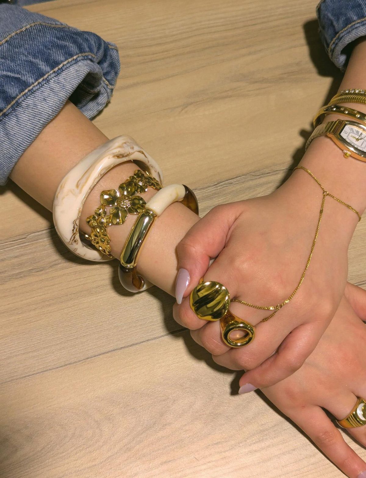 19 Jewelry Aesthetic Gold Ideas for Easy Style 7 Hands wearing gold rings, bangles, and a chain bracelet on a wooden table.